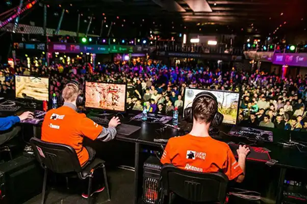 Dva hráči v oranžových tričkách a se sluchátky hrají počítačové hry na pódiu během esportového turnaje, zatímco v pozadí je sleduje velké množství diváků.